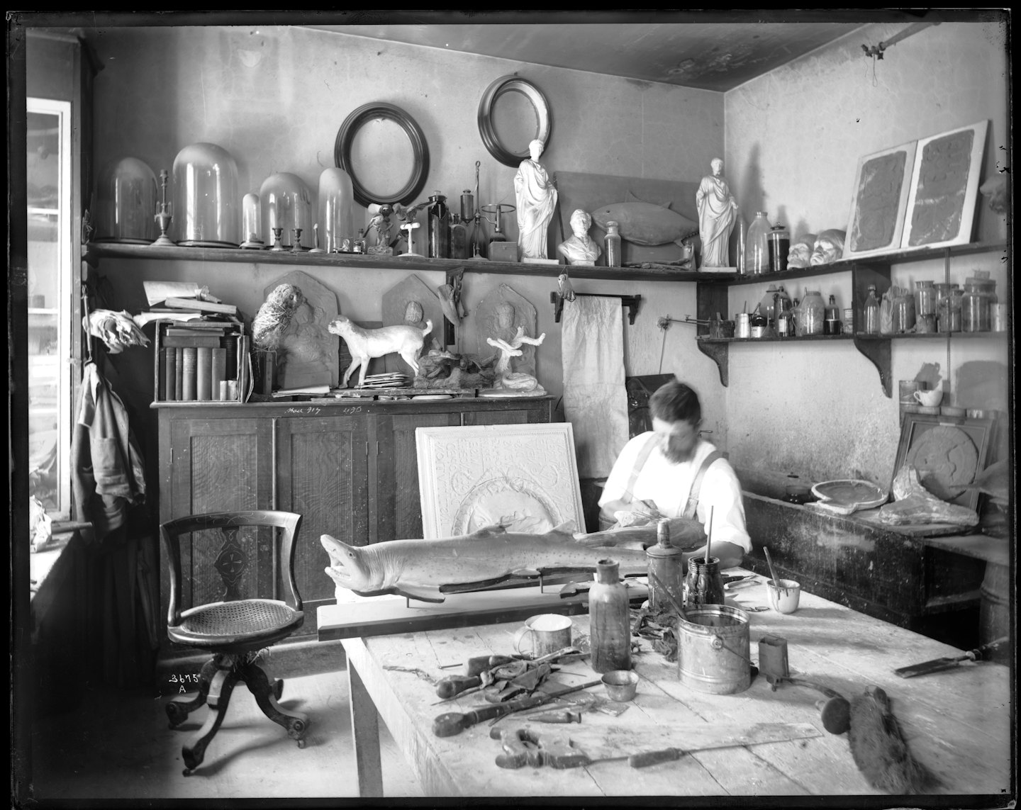 Artisan carves a large fish model in a cluttered workshop.